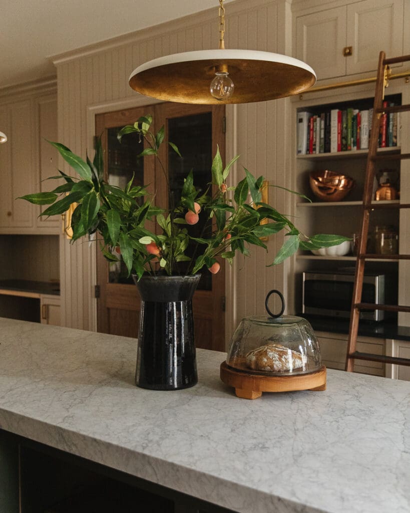 Cloche with baked bread underneath on a kitchen countertop next to a large vase with springs of peaches