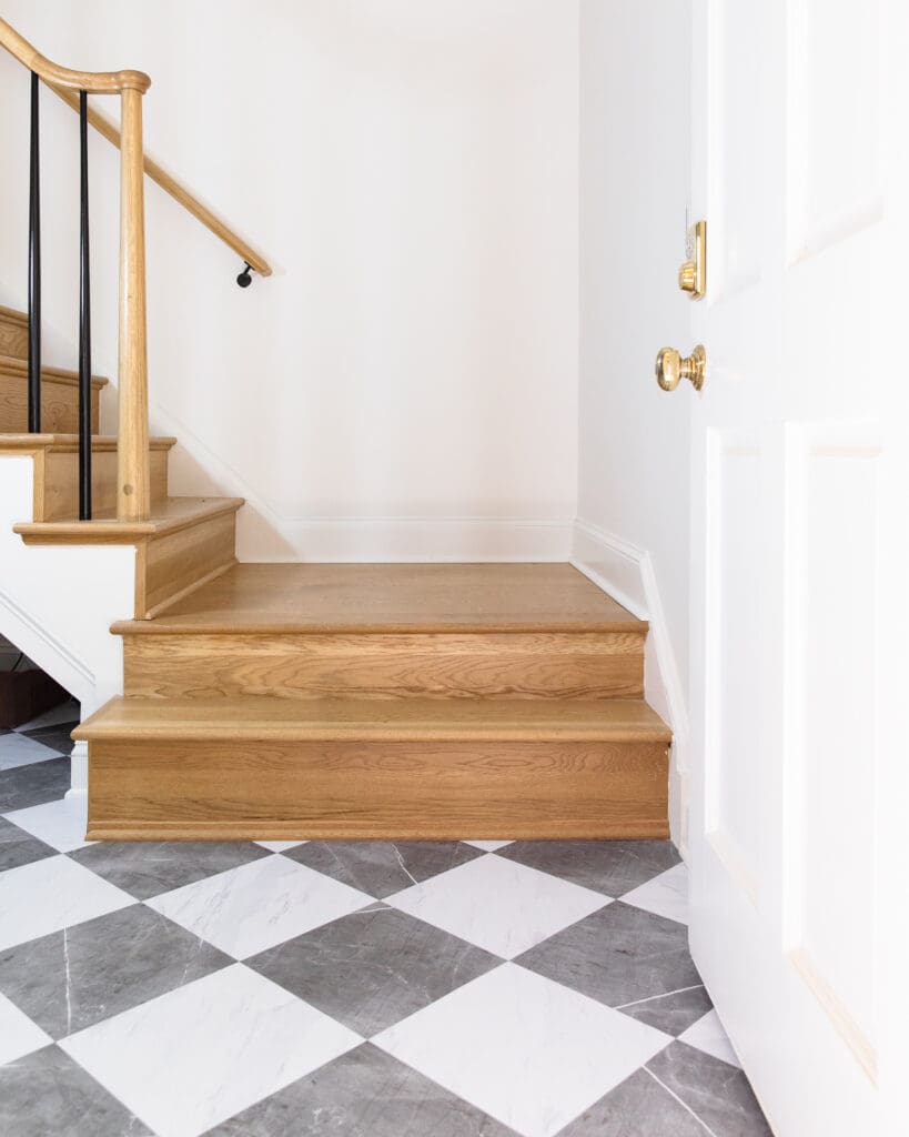 Elegant staircase with natural wood steps and a simple black metal handrail, set against a white wall with a patterned tile floor.