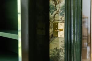 Up of a brass door lock and green-painted door frame with decorative wallpaper in the background.