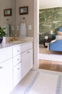 An inviting bathroom with white cabinetry and gold fixtures, seamlessly connected to a stylish living room featuring a blue armchair and nature-inspired wallpaper.