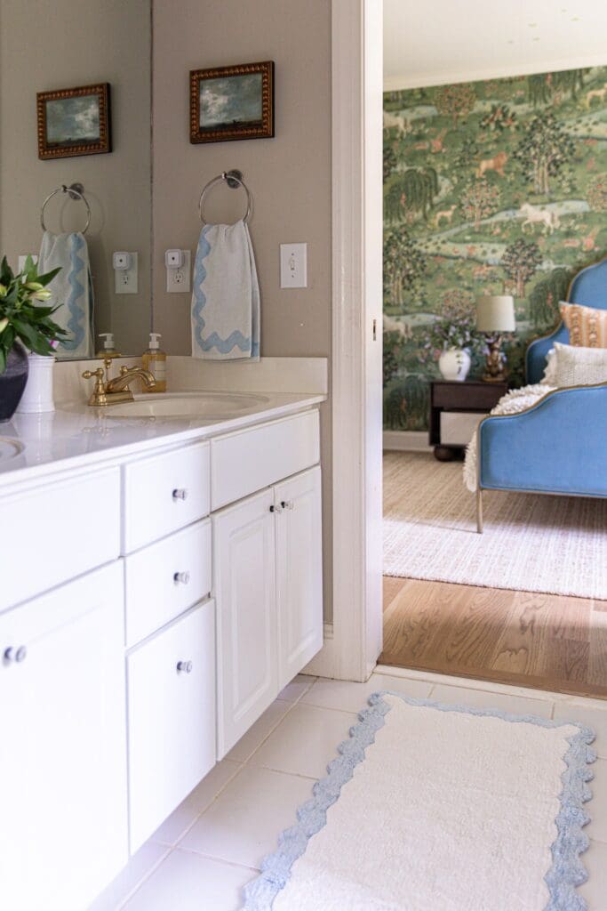 An inviting bathroom with white cabinetry and gold fixtures, seamlessly connected to a stylish living room featuring a blue armchair and nature-inspired wallpaper.
