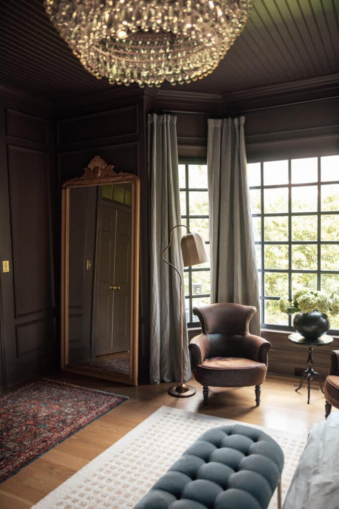A beautifully styled bedroom featuring dark wall paneling, classic armchairs, a large window with curtains, a decorative mirror, and a chandelier, showcasing elegant home decor