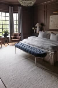 Cozy bedroom featuring large windows, elegant drapes, a plush bed with white linens, a velvet bench at the foot of the bed, and vintage-style furniture creating a warm, inviting atmosphere.