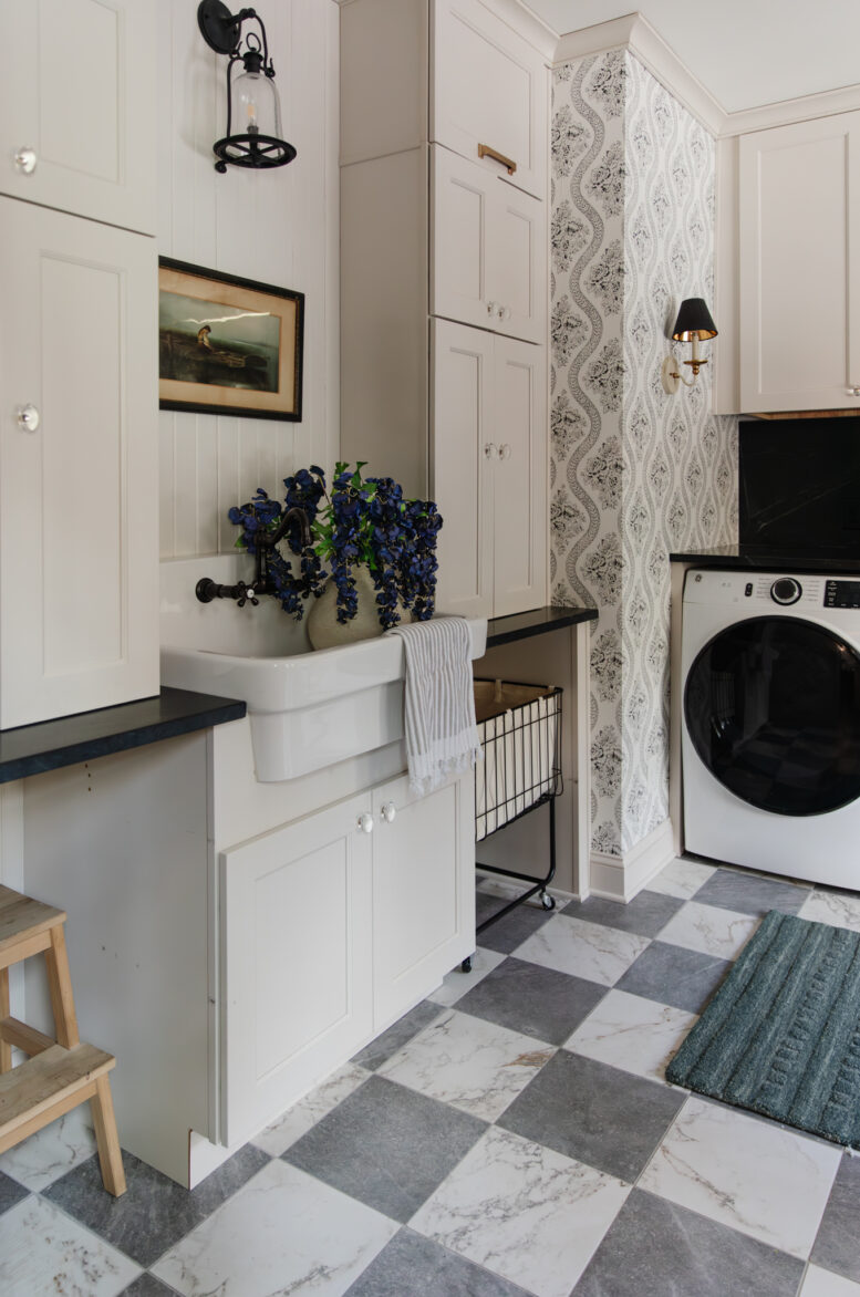 A laundry room with cream cabinets, black and white wallpaper, gray and white marble checker floors, and a porcelain sink full of a vase of purple flowers.