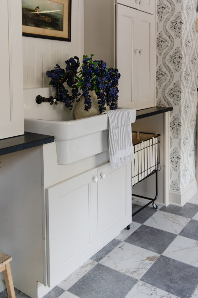 A laundry room with cream cabinets, black and white wallpaper, gray and white marble checker floors, and a porcelain sink full of a vase of purple flowers.