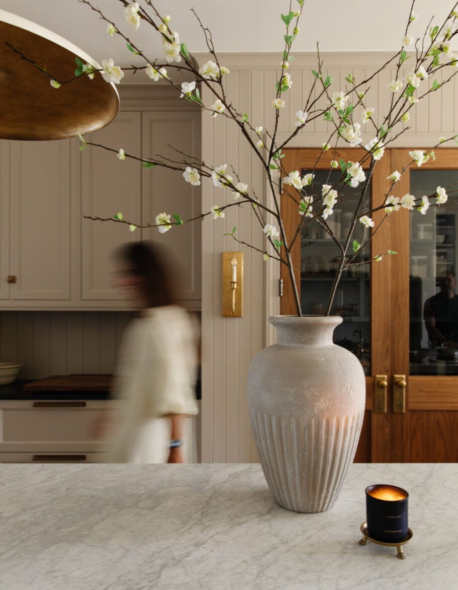 A woman walking past a kitchen island with marble countertops, a ceramic vase on the counter filled with cherry blossom stems and a lit candle on a brass candle pedestal.
