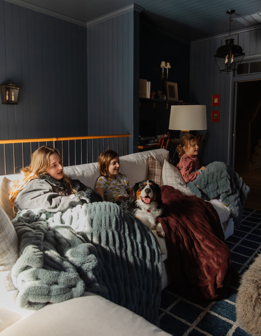 Family enjoying a cozy movie night on the sofa with their dogs, surrounded by blankets and warm lighting in a stylish living room. Perfect for family movie time and relaxing evenings.