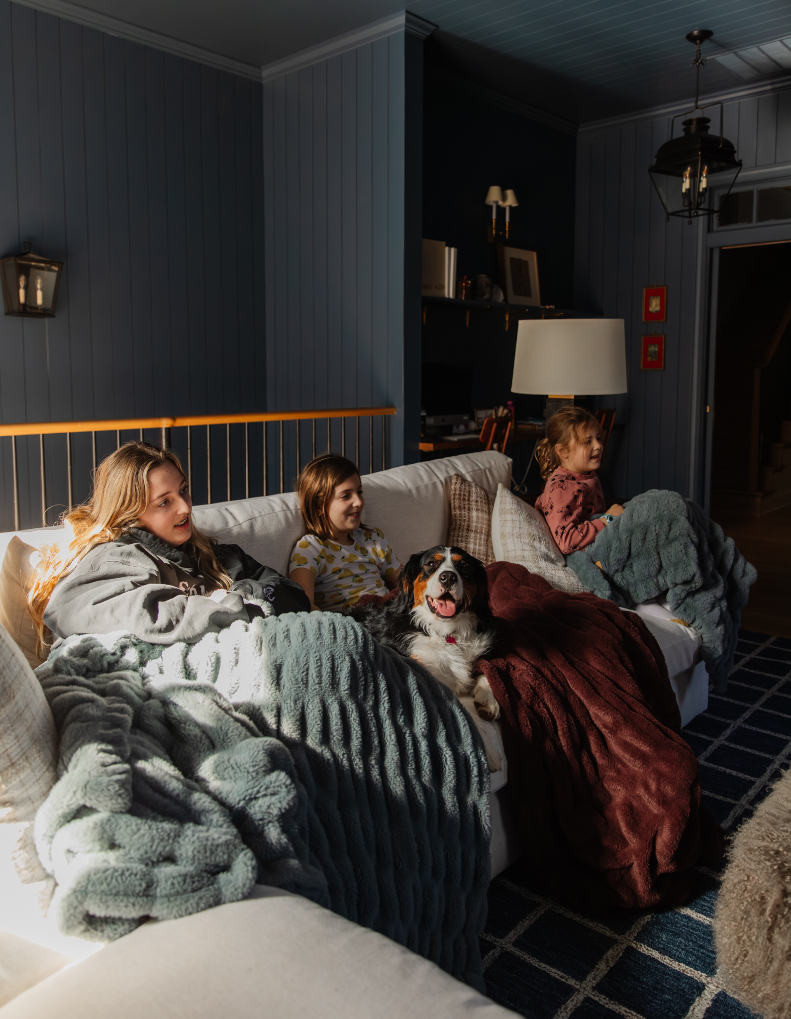 Family enjoying a cozy movie night on the sofa with their dogs, surrounded by blankets and warm lighting in a stylish living room. Perfect for family movie time and relaxing evenings.