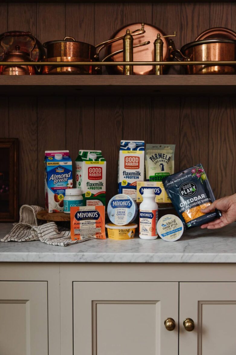 Cream cheese and dairy-substitute products on kitchen countertop with copper pots on wooden shelf above.