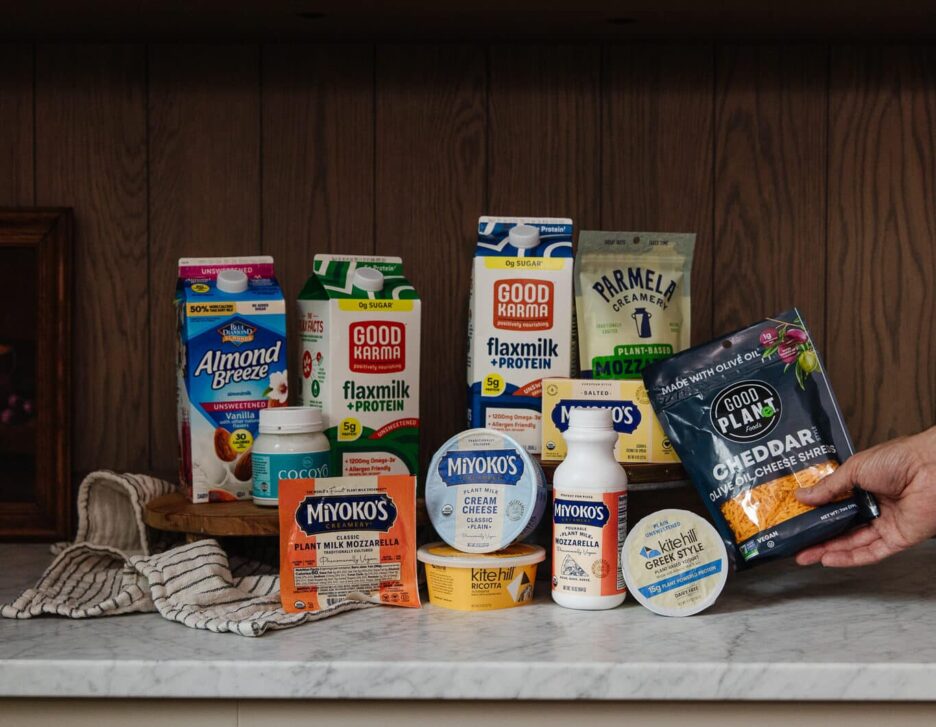 A variety of plant-based milk, cheese, and yogurt products from brands like Miyoko's, Good Karma, and Parmela, displayed on a kitchen counter with a hand holding a cheese wedge.