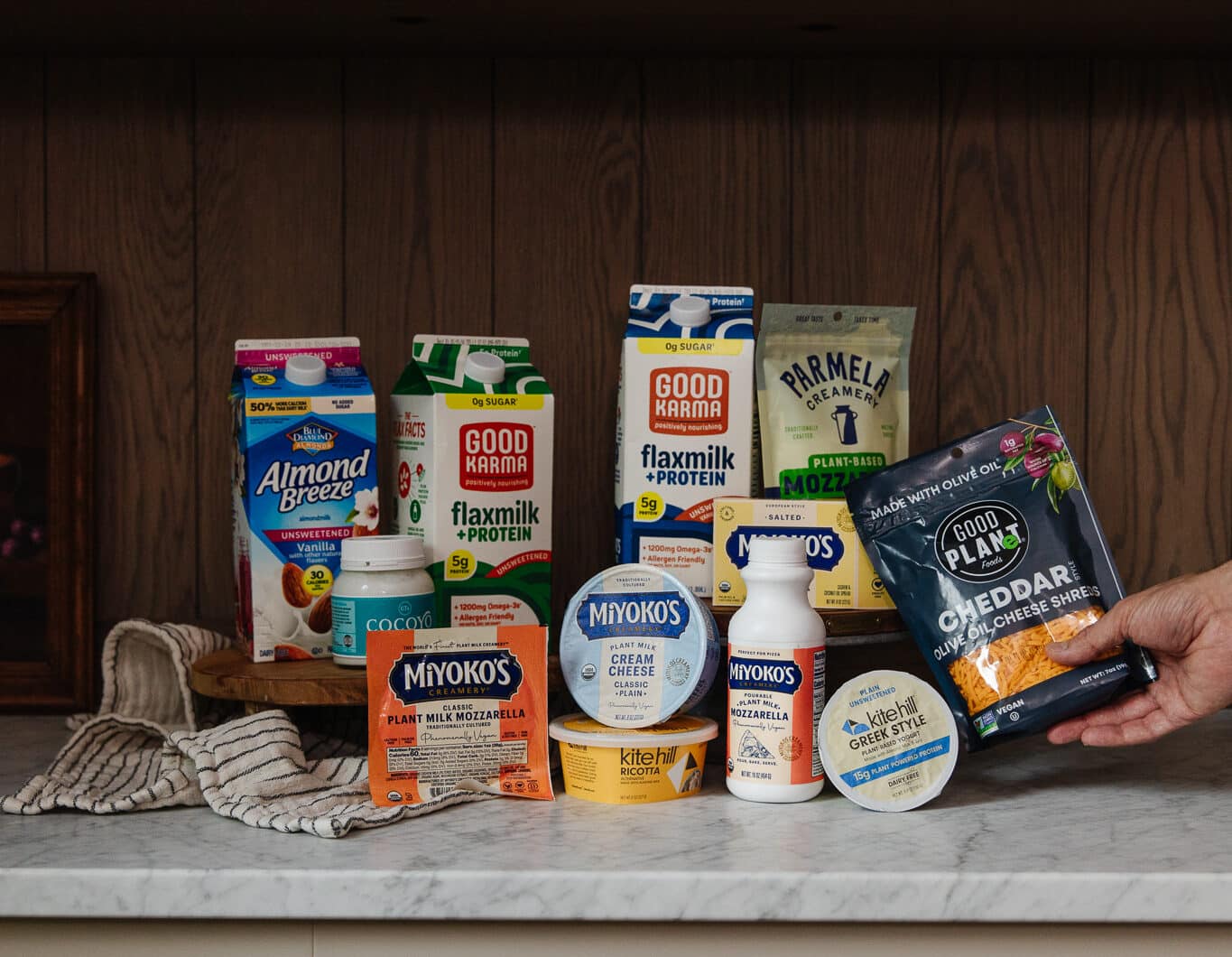 A variety of plant-based milk, cheese, and yogurt products from brands like Miyoko's, Good Karma, and Parmela, displayed on a kitchen counter with a hand holding a cheese wedge.
