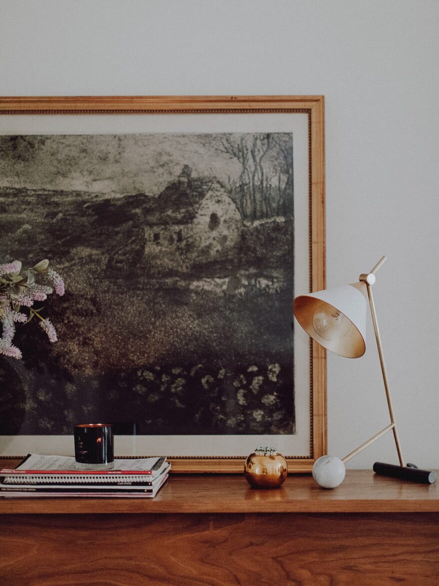 Charming wall art decor enhances living space with rustic elegance, complemented by modern accessories like a sleek table lamp, flowers, and neatly arranged books for a warm, inviting look.