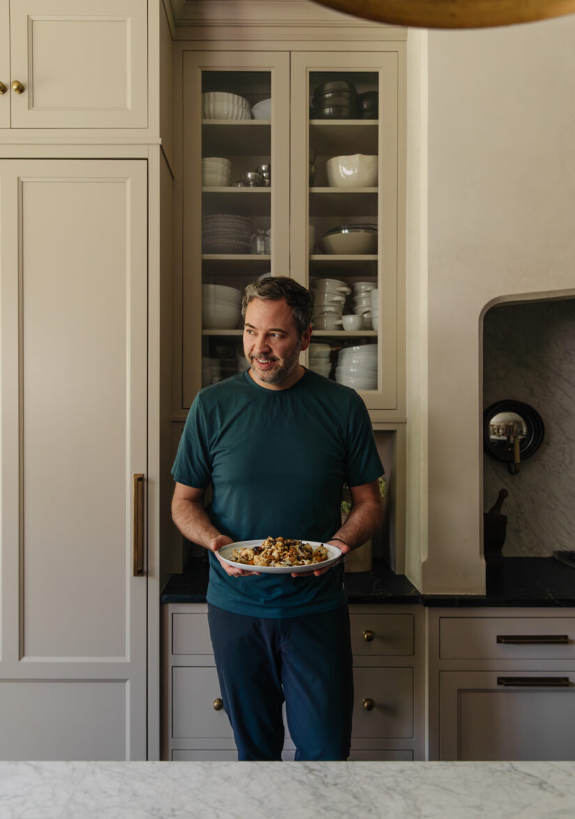 A man stands in a modern kitchen holding a plate of roasted cauliflower drizzled with homemade dairy-free Caesar dressing, highlighting a delicious and healthy plant-based dish.