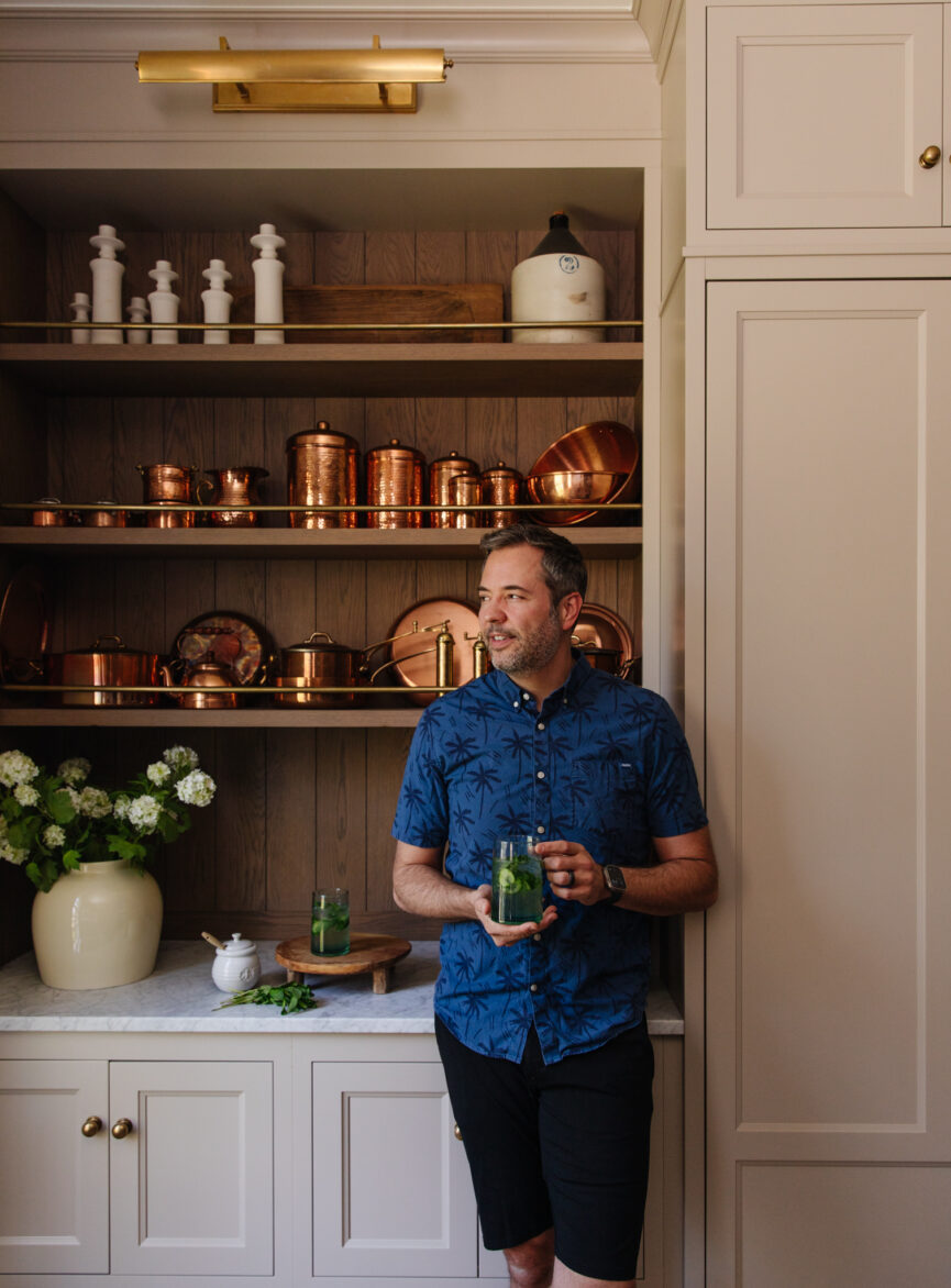 Chris Marcum holding a cucumber honey "Nojito" cocktail in a bright kitchen with copper cookware and white cabinets.