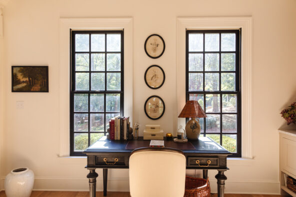 A cozy home office with a black desk between two large windows, decorated with books, a lamp, and office supplies, creating an organized workspace.