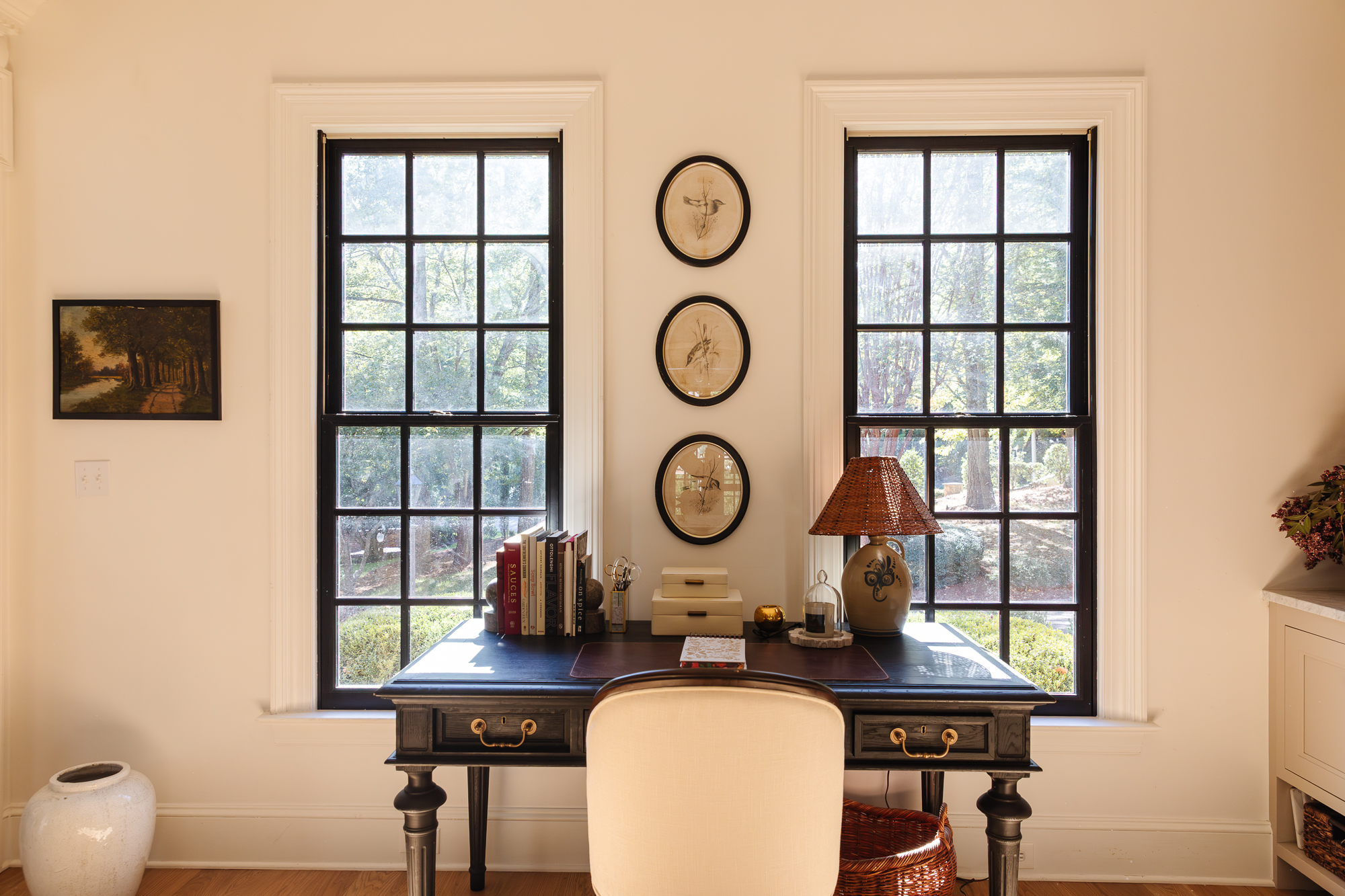 A cozy home office with a black desk between two large windows, decorated with books, a lamp, and office supplies, creating an organized workspace.
