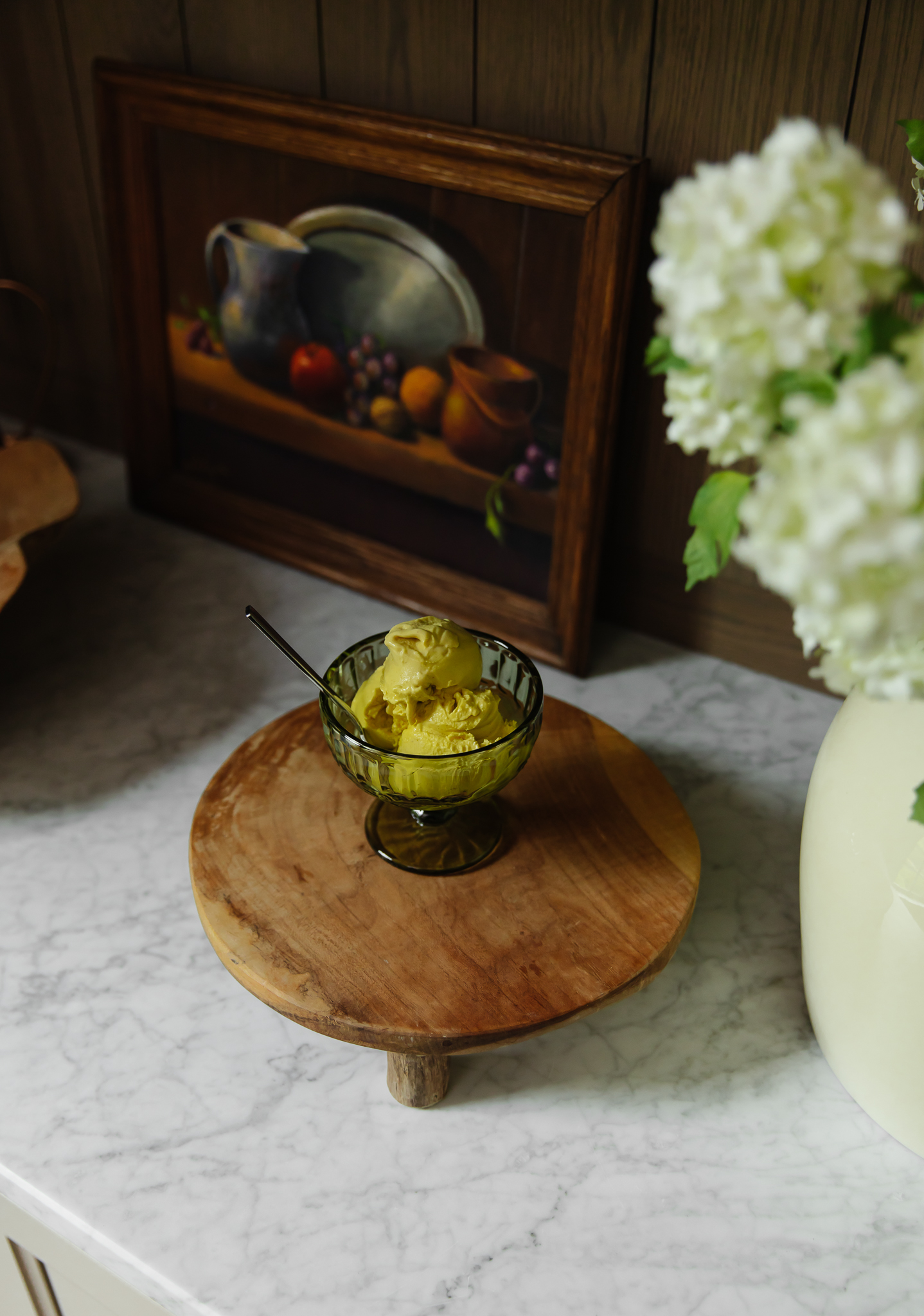 Dairy-free golden milk ice cream served in a glass bowl on a wooden stand, with a framed still life painting and white hydrangeas in the background, perfect for a healthy dessert.