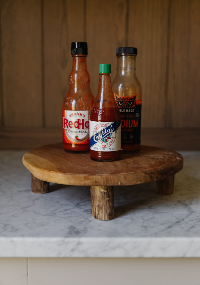 Frank's Red Hot, Crystal Hot Sauce, Noble Made Medium Buffalo Sauce displayed on a small wooden stand on a marble countertop, showcasing popular condiments for spicy food lovers.