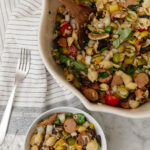 A colorful vegetable pasta salad featuring cherry tomatoes, sliced sausage, celery, and pasta, served in a white bowl on a marble surface.