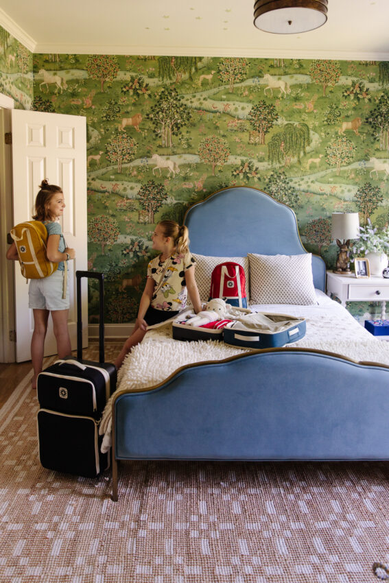 Children preparing luggage in a colorful, inviting bedroom with a nature-inspired mural and a blue upholstered bed.