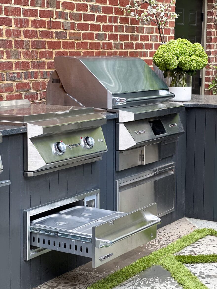 Modern stainless steel outdoor grill and oven with side drawer, set against a brick wall, featuring a potted green plant for a stylish outdoor cooking area.