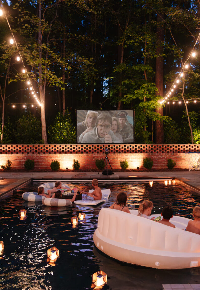 Outdoor movie night setup with a projector screen and people on pool floats in a luxury backyard pool.