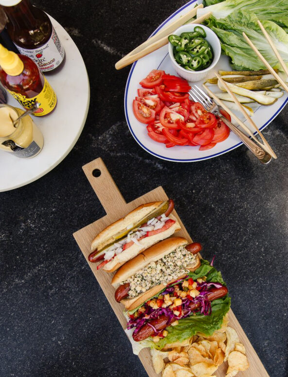 Delicious fresh tomato salad with sliced tomatoes, served alongside hot dogs and a variety of sandwiches on a dark countertop. Perfect for summer meals or casual gatherings.
