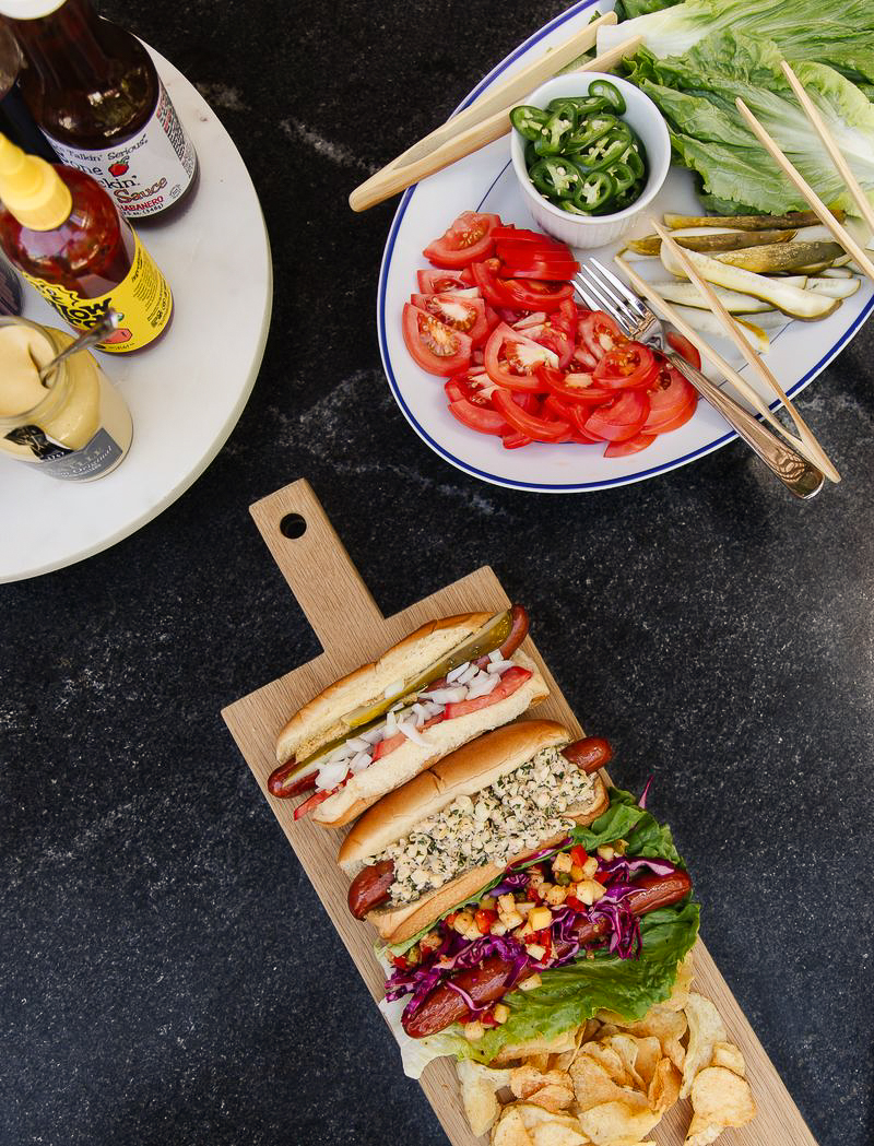 Delicious fresh tomato salad with sliced tomatoes, served alongside hot dogs and a variety of sandwiches on a dark countertop. Perfect for summer meals or casual gatherings.