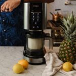 Ninja CREAMi ice cream maker with digital touchscreen interface on a modern kitchen countertop, surrounded by fresh pineapple and lemons, showcasing a stylish and functional kitchen appliance.