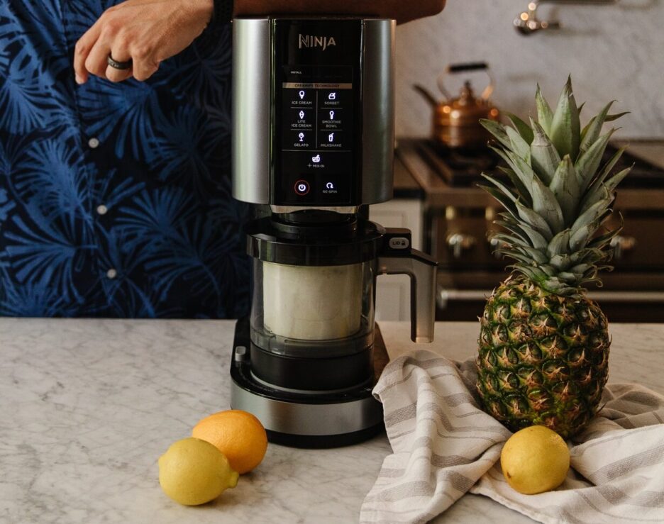 Ninja CREAMi ice cream maker with digital touchscreen interface on a modern kitchen countertop, surrounded by fresh pineapple and lemons, showcasing a stylish and functional kitchen appliance.