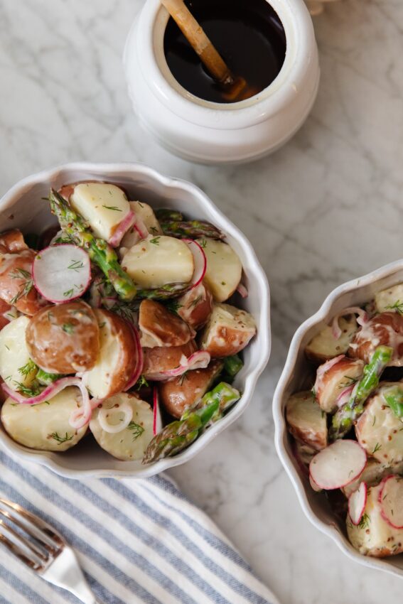 Delicious potato salad featuring radishes, asparagus, and a creamy dressing, served in white bowls on a marble surface. Perfect for summer gatherings or casual meals.