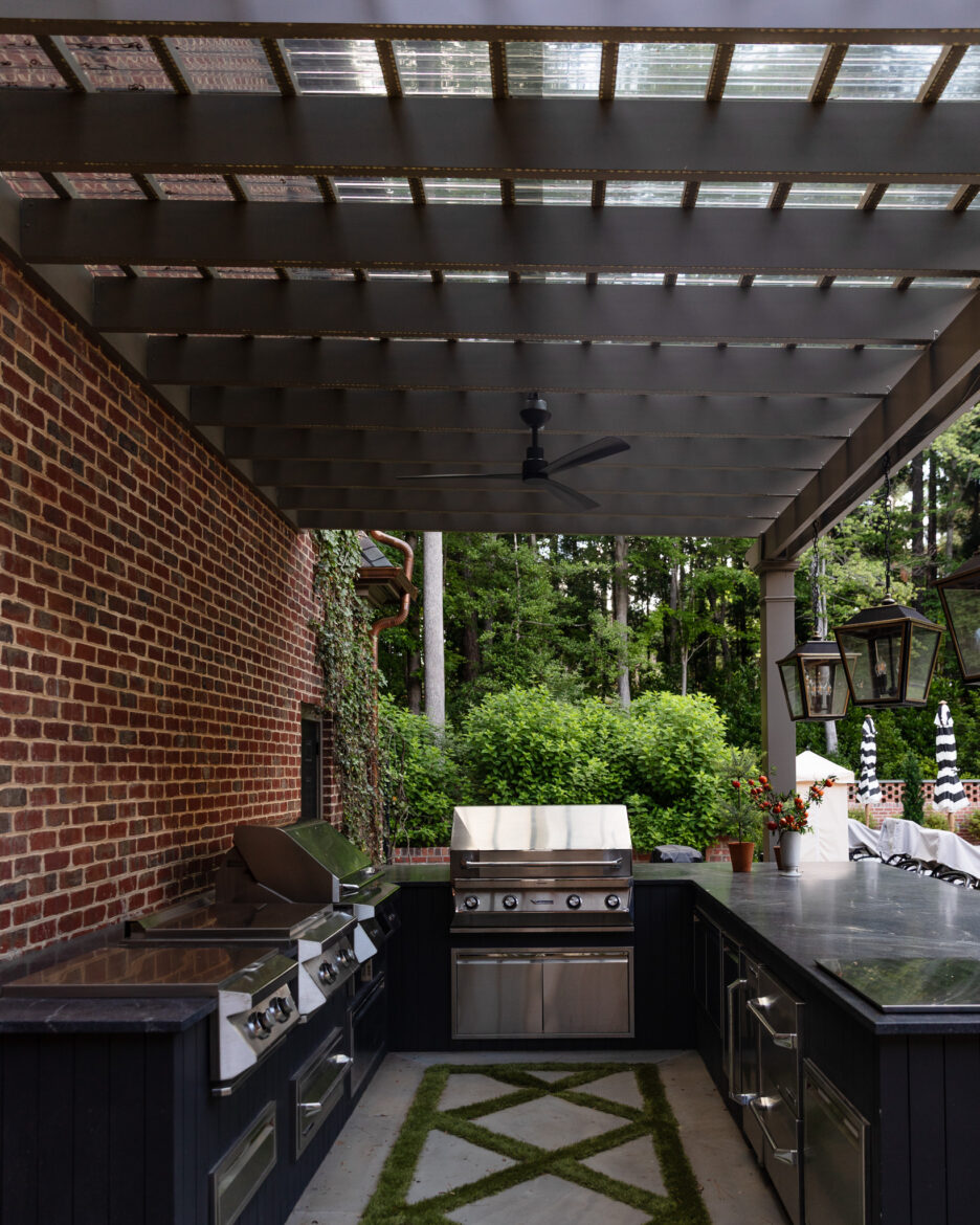 Outdoor covered kitchen area featuring a brick wall, stainless steel grill, black cabinetry, and lush green plants in the background, perfect for outdoor cooking and entertaining.