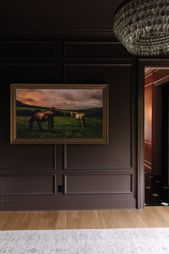 Elegant framed horse artwork in a stylish living room with dark paneled walls and a crystal chandelier overhead.