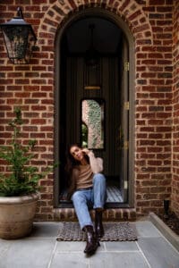 Julia sitting on the mudroom stoop wearing a brown top, denim jeans, and brown leather boots
