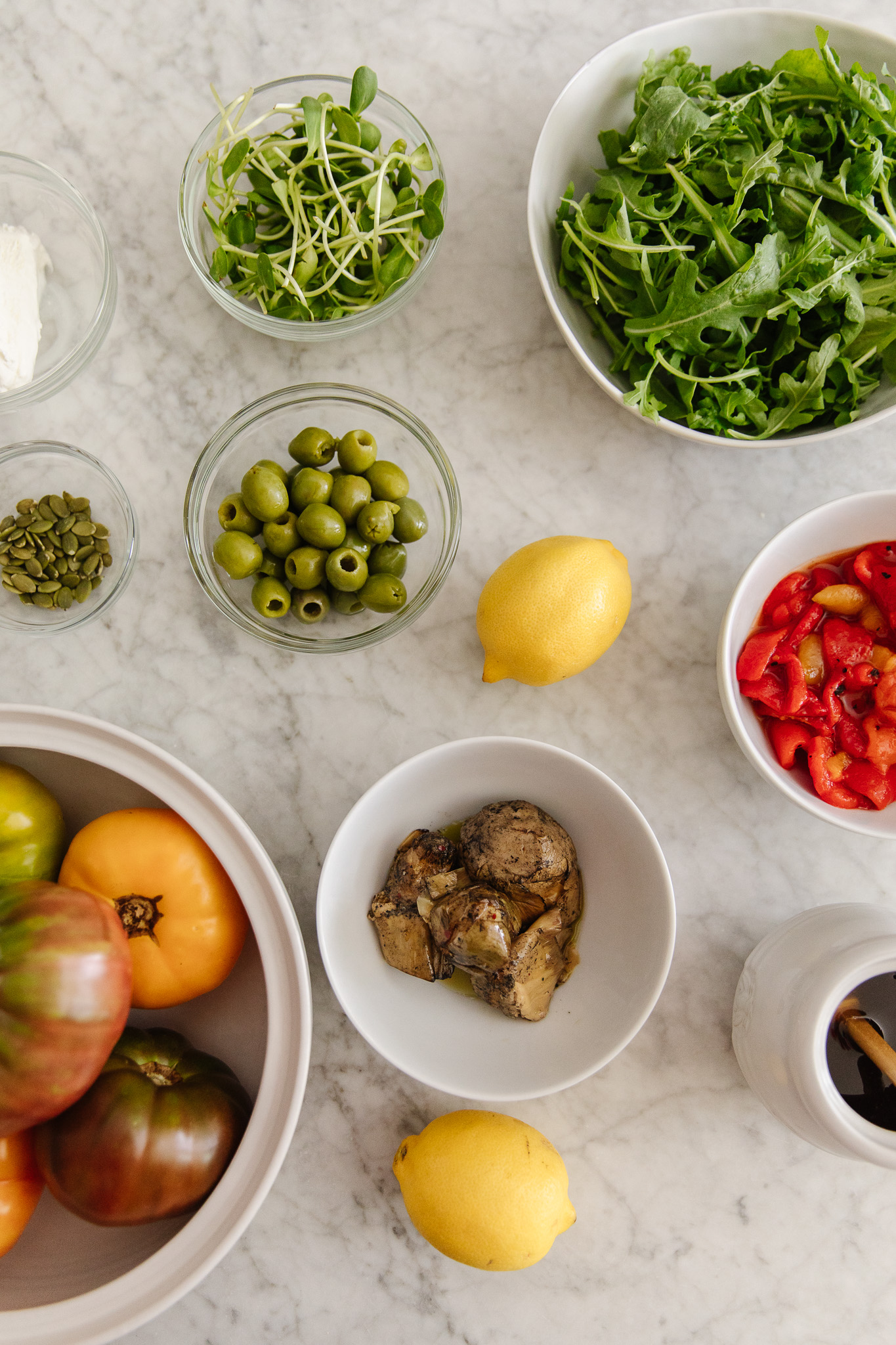 Chris Loves Julia | Flatlay of ingredients for the tomato salad including roasted red peppers, marinated artichoke hearts, honey, lemon, olives, pumpkin seeds and arugula