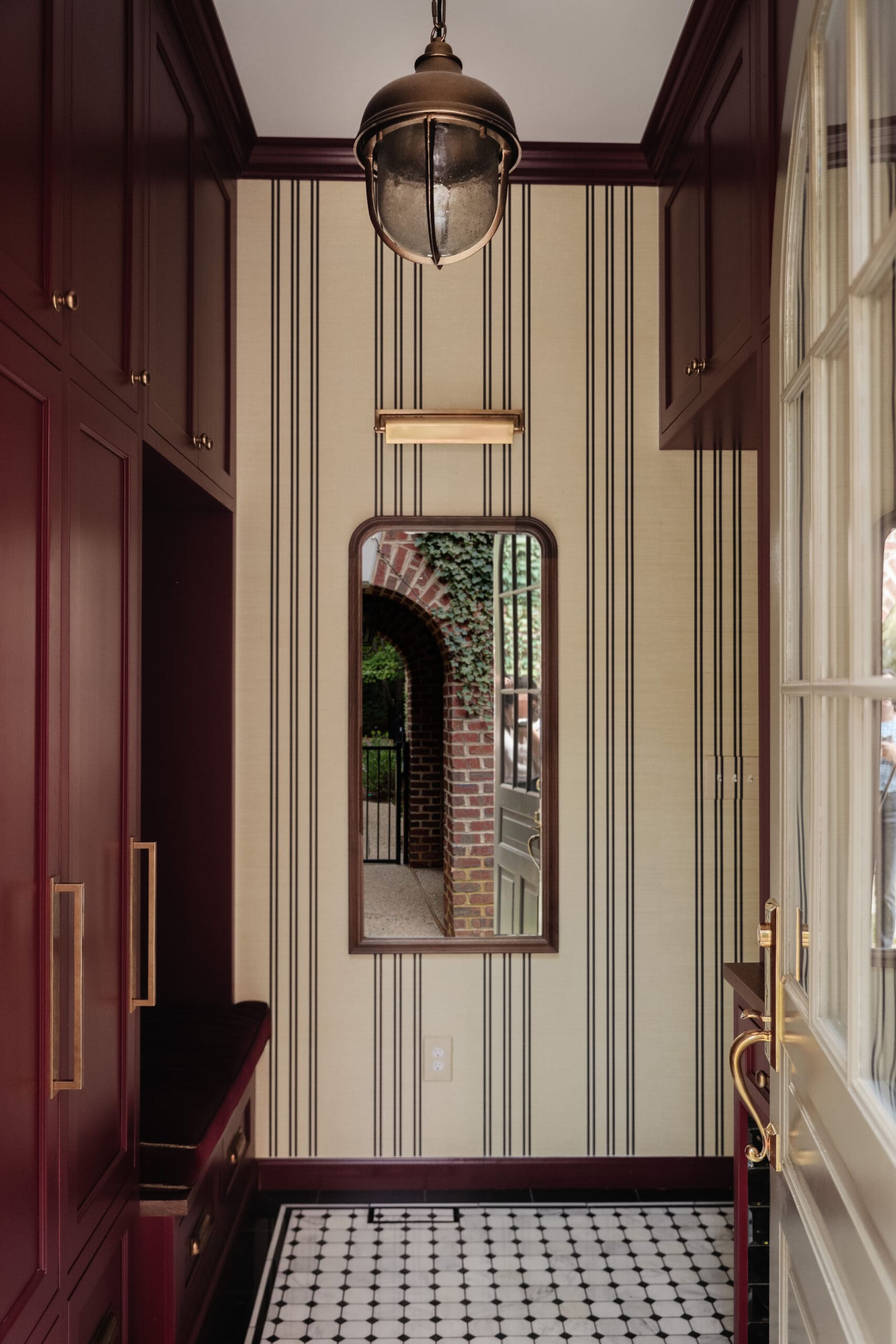 Chris Loves Julia | Moody dark red mudroom with grasscloth striped wallpaper and a mirror