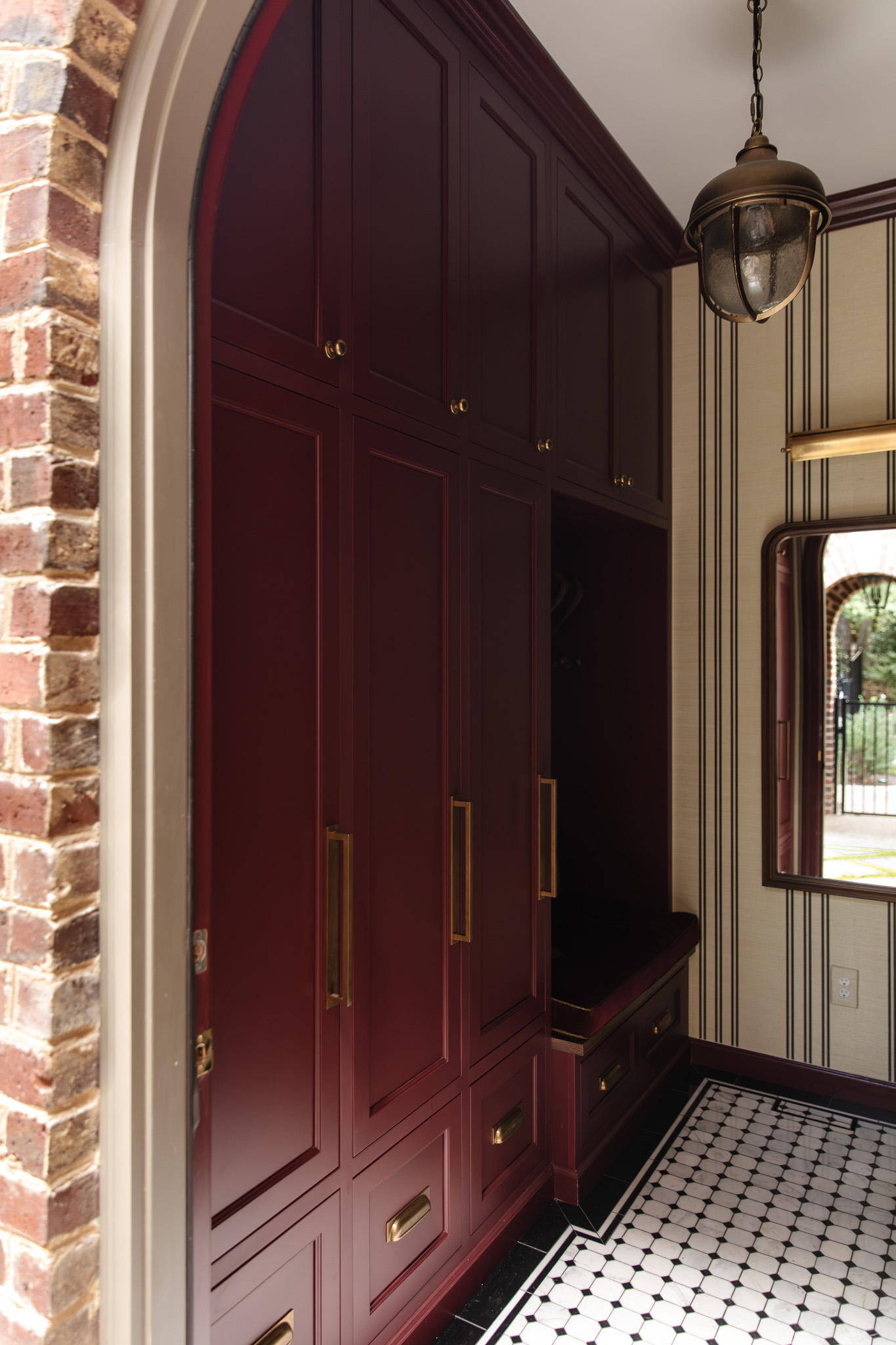 Chris Loves Julia | Moody dark red mudroom with gold locker accents and a built-in bench seating area