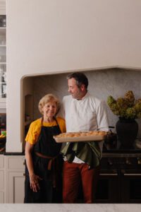 Chris in the kitchen holding a baking pan of dinner rolls next to his mom