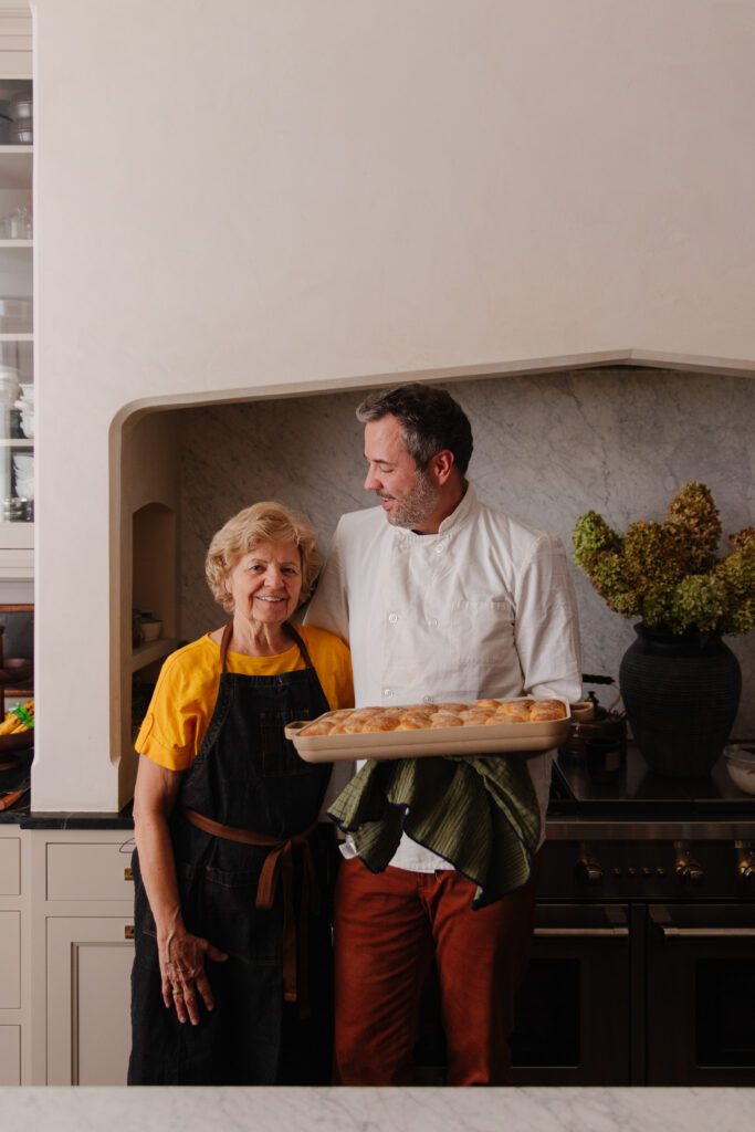 Chris in the kitchen holding a baking pan of dinner rolls next to his mom