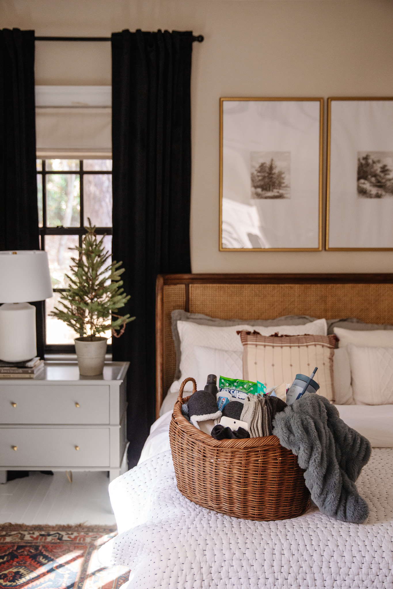 Basket of miscellaneous items on a cozy bedroom bed with layered white bedding, next to a white nightstand with a small potted houseplant, near a window with dark curtains, in a modern rustic bedroom.