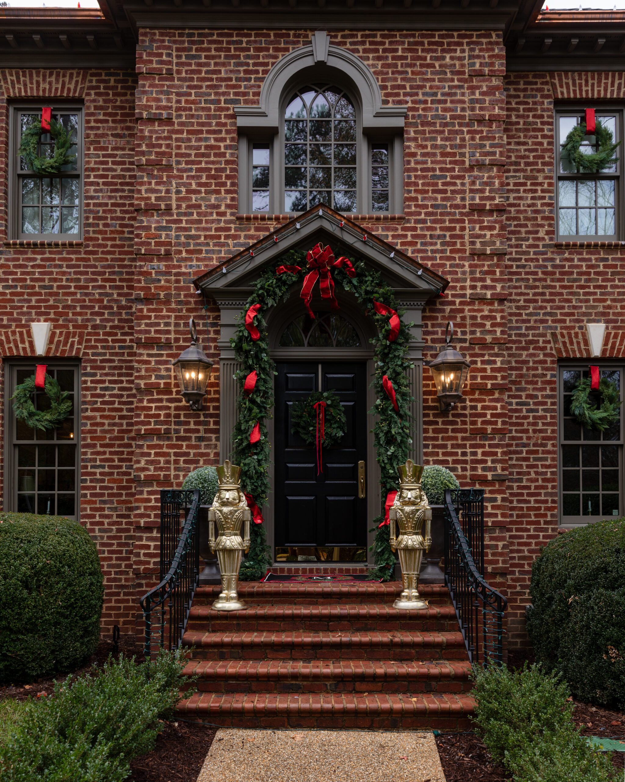 The front door of a brick Colonial house with tall gold nutcrackers and a draping green and red garland around the door