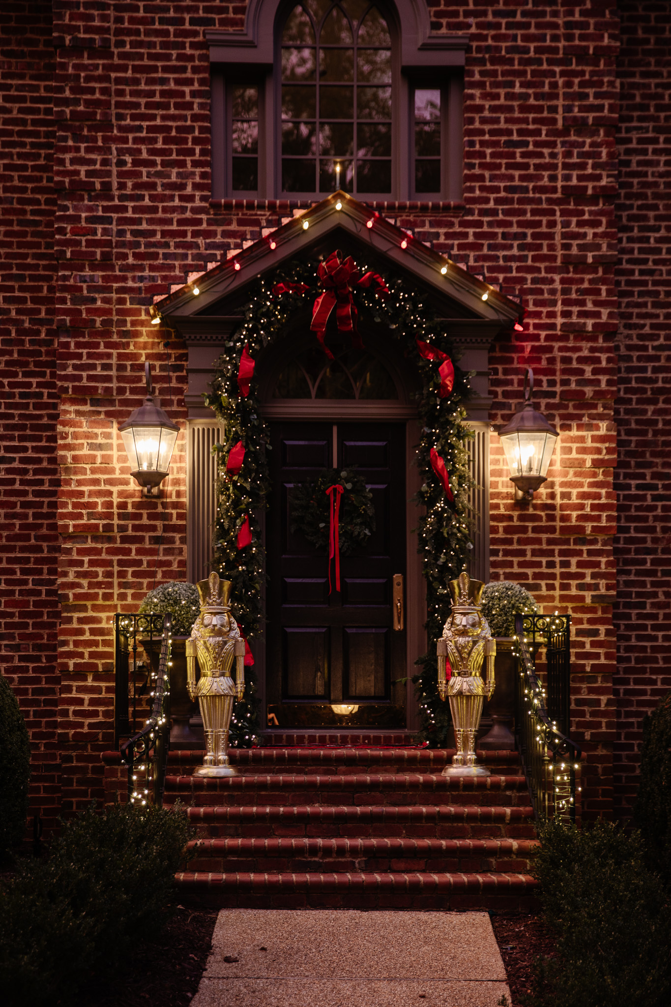 A front door and stoop at dusk illuminated by Christmas lights and decorated with a red-and-green garland and gold nutcrackers