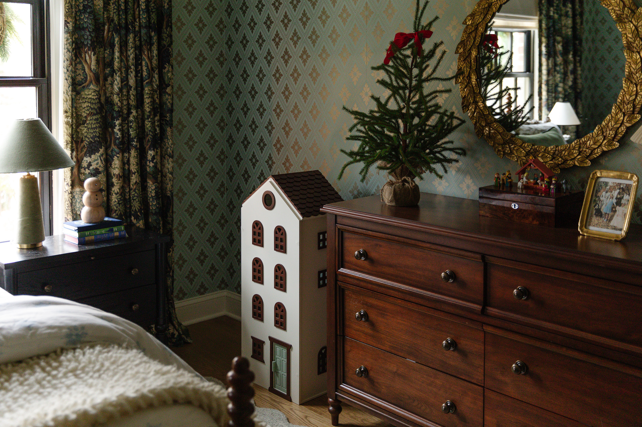 A girl's bedroom dresser topped with a small spruce tree topped with a single red bow and a tall dollhouse in the corner