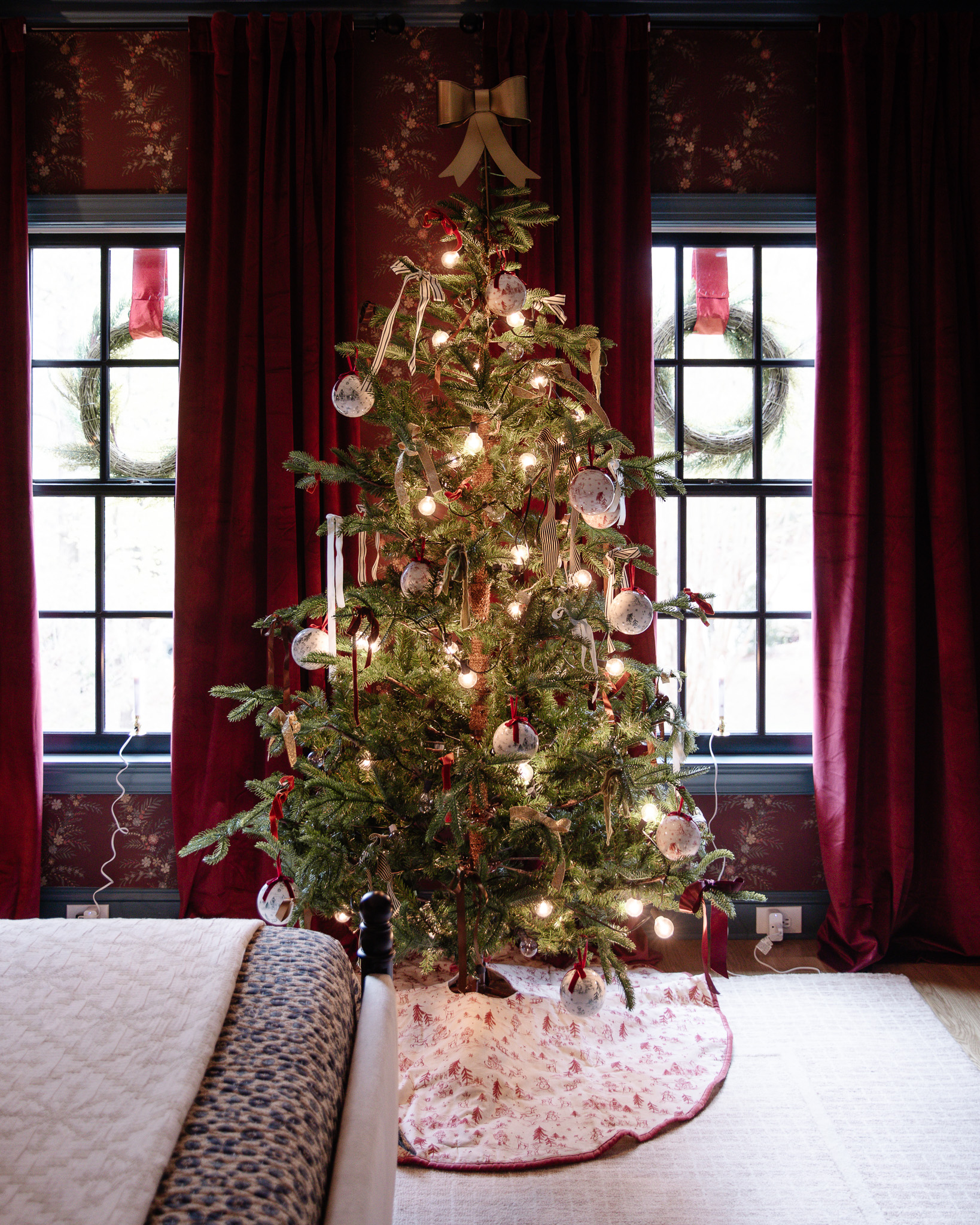 A traditional-style Christmas tree with toile baubles and red bows in a burgundy red bedroom.