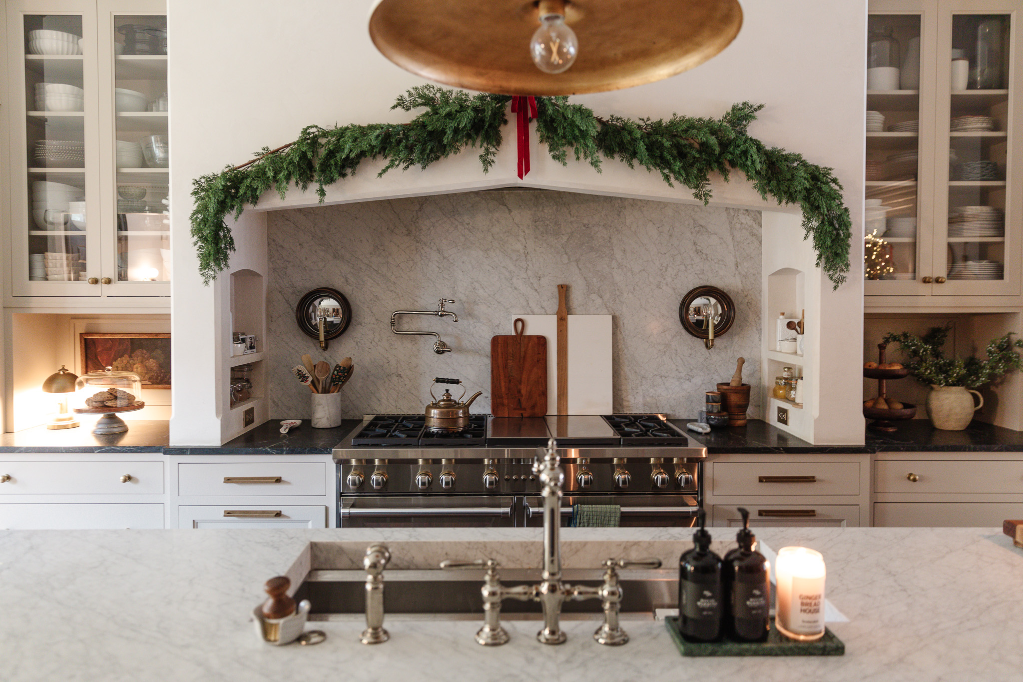 A white kitchen with marble countertops and backsplash and a green garland over the hood with a red bow