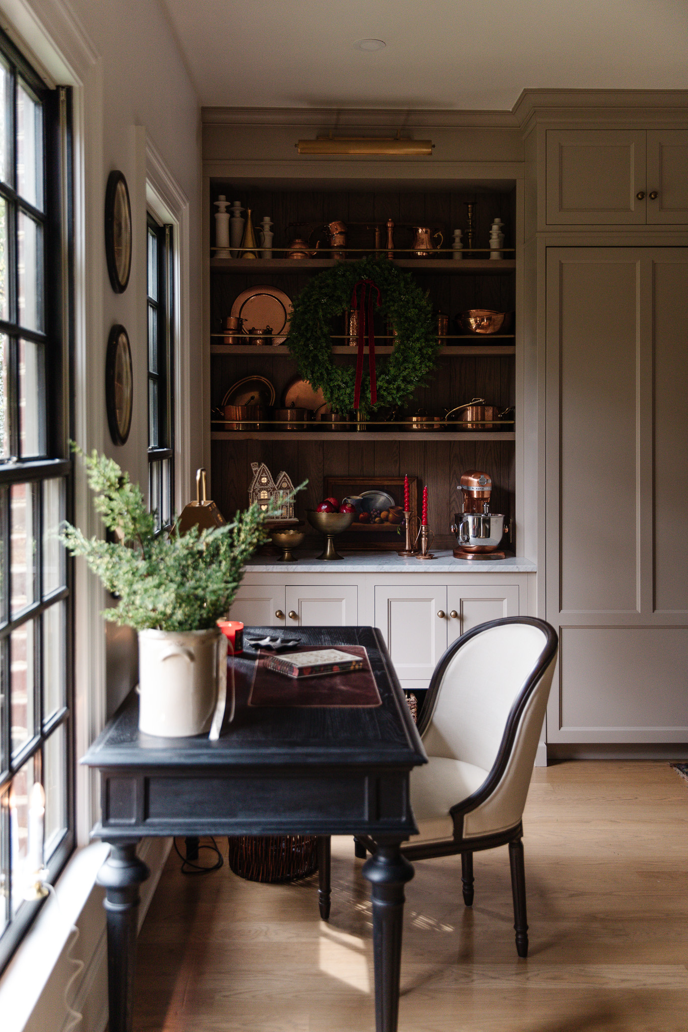 A kitchen desk and shelves decorated for Christmas with red twirl candles, a large wreath on the open shelves, and an illuminated ceramic gingerbread house