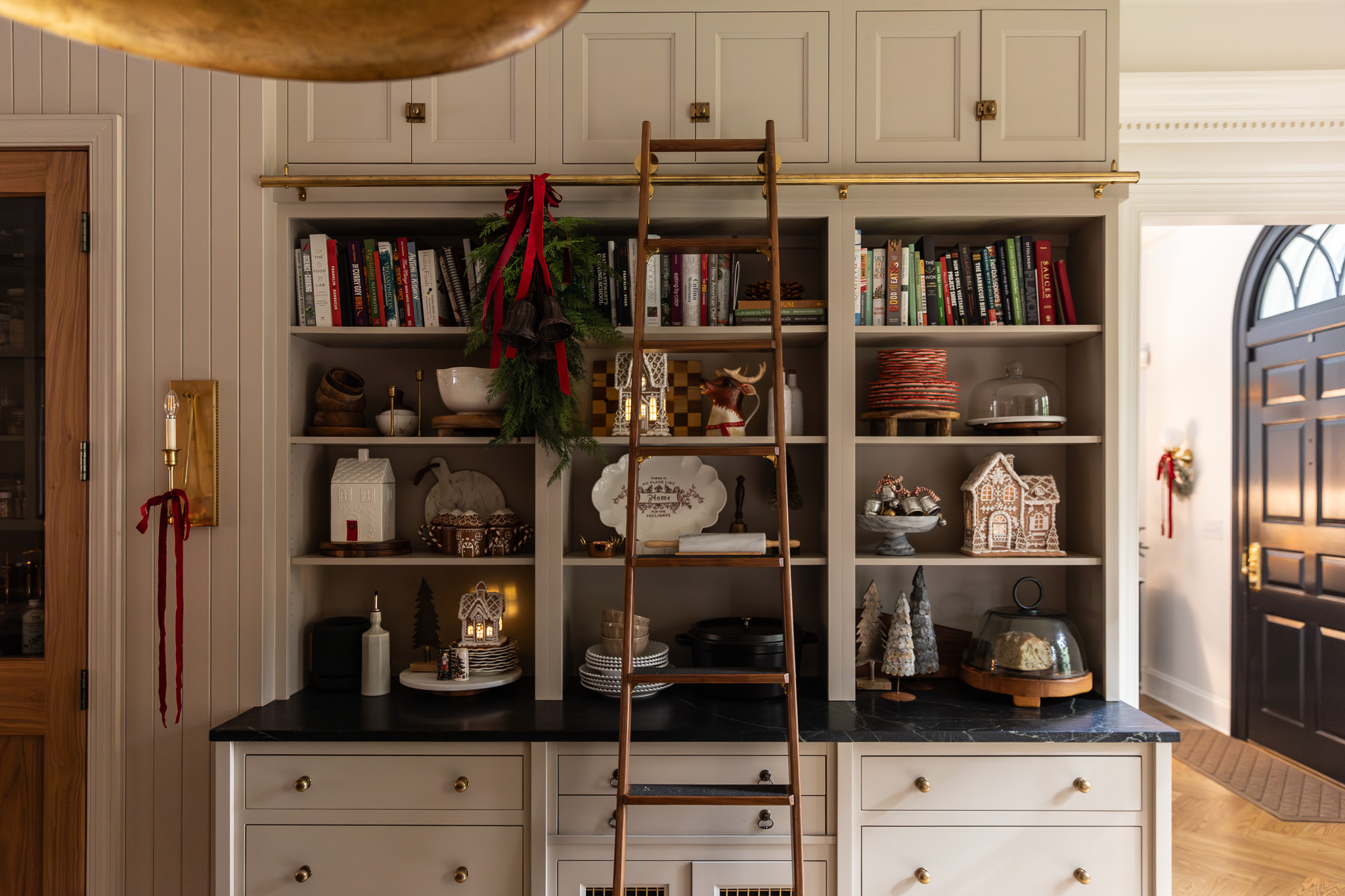 Open kitchen shelves with Christmas decorations including ceramic gingerbread houses, gingerbread house mugs, a greenery swag with red ribbon and bells, and red plates