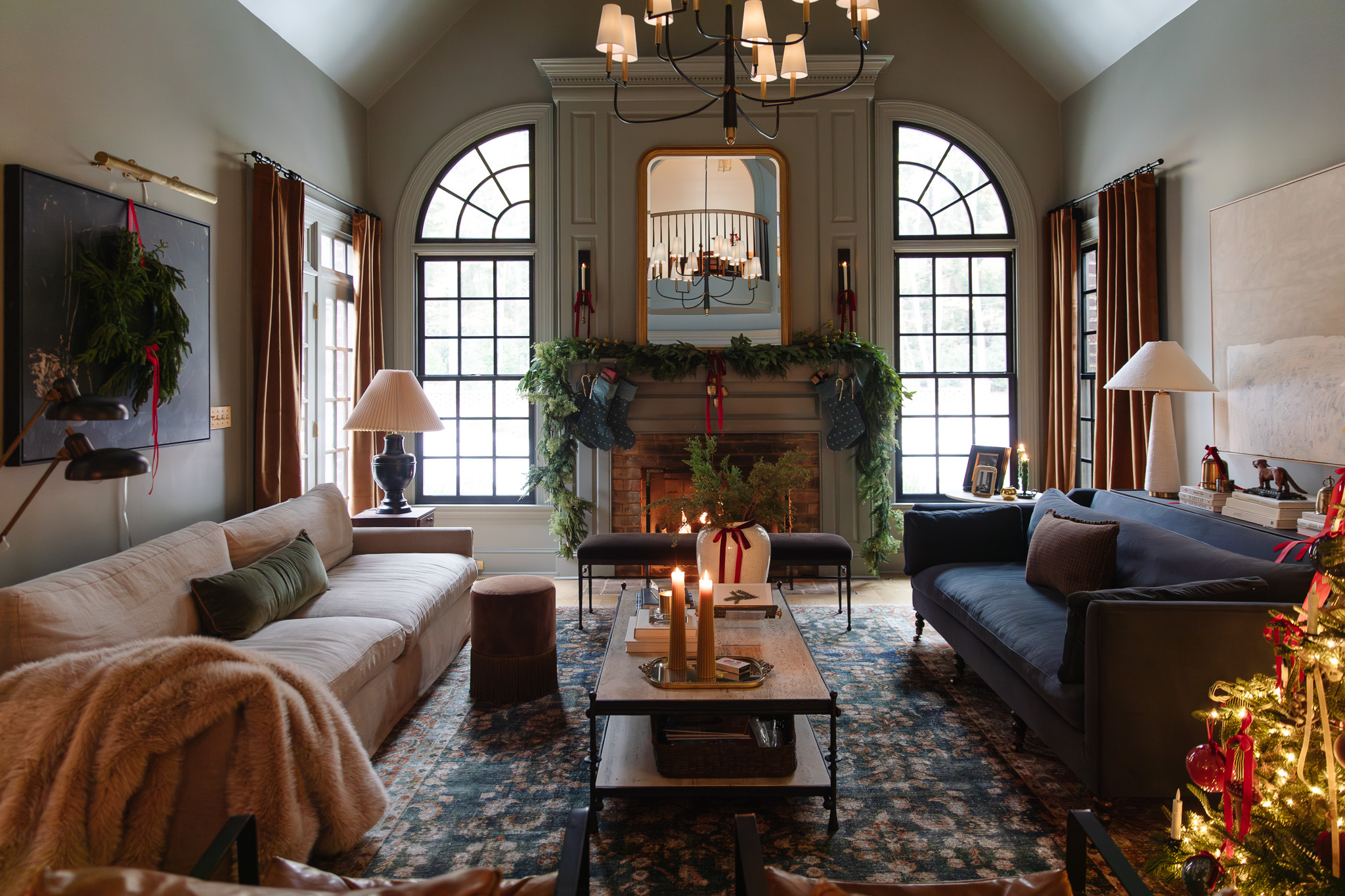 A warm, cozy traditional living room with a lush green garland on the mantle, a wreath on the wall, green stockings, and lit candles.