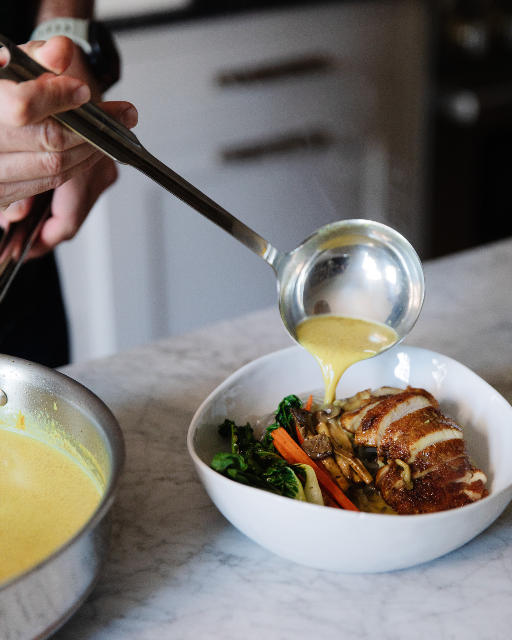 A ladle pouring Singapore-style coconut curry broth into a soup bowl filled with veggies and chicken