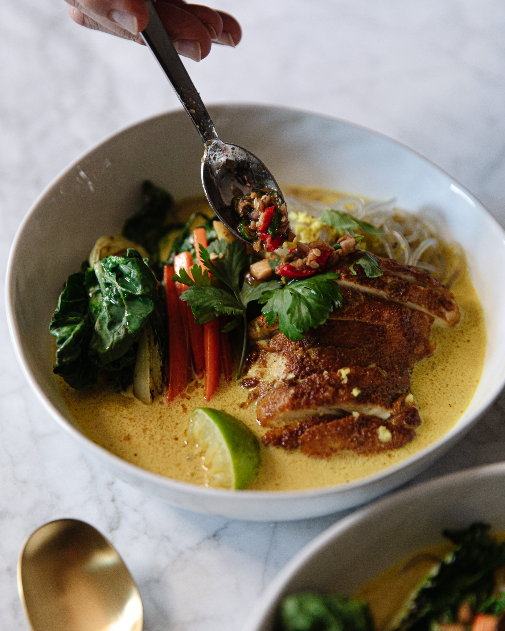 A soup spoon drizzling sweet & spicy chili oil into a Singapore Noodles-inspired Chicken Noodle Soup